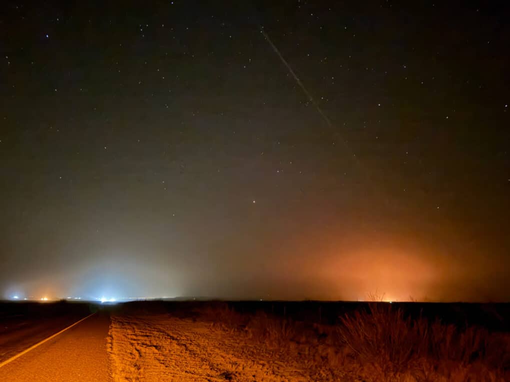 Color photograph of a dark sky at night, with a low horizon on the shoulder of a road, with glow red and white lights glowing in the distance.