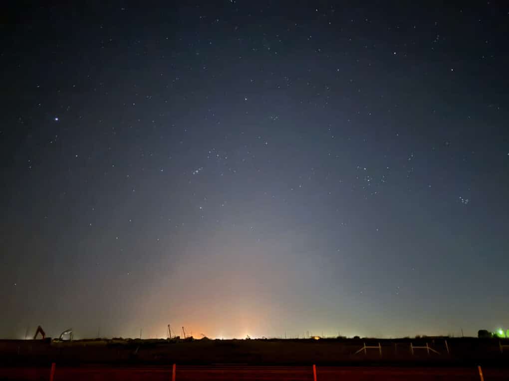 Color photograph of a dark sky at night, with a low horizon against which glow red and green lights.