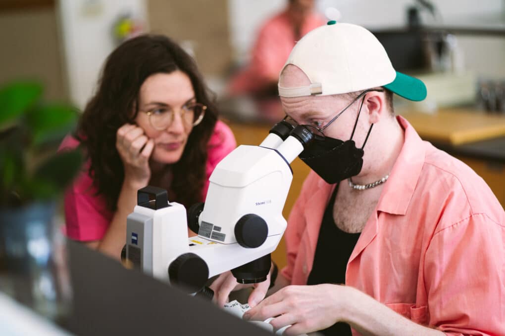 One person is looking into a microscope, wearing a facemask, a backwards baseball cap, and a pink lab coat. Another person, immediately beside, is watching. 