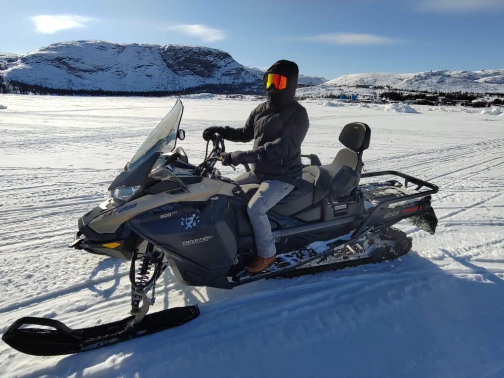 Snowy landscape in background, and in the foreground a jet ski-type device designed to move on ice rather than water. The machine has two seats, and the driver is extremely bundled up for severe cold, with no skin visible. 