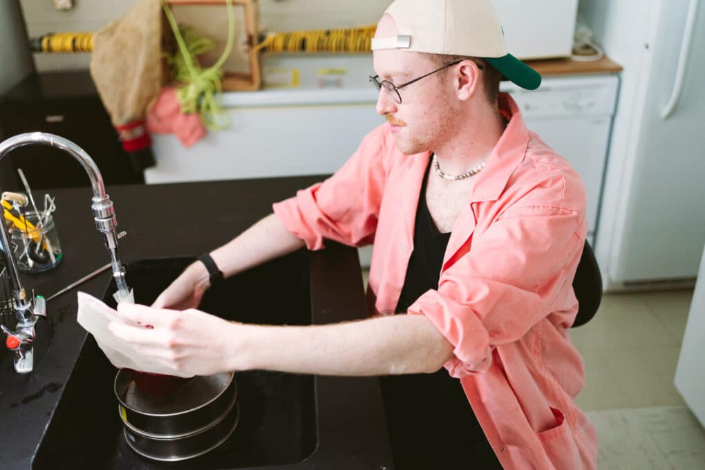 A man wearing a baseball cap backwards and glasses and pink lab coat is rinsing a small fine mesh funnel in a sink. 