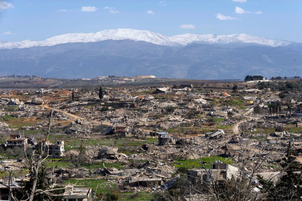 Wide-angle color photograph of small buildings reduced to rubble in a rural area, with snow-capped mountains in the distance.