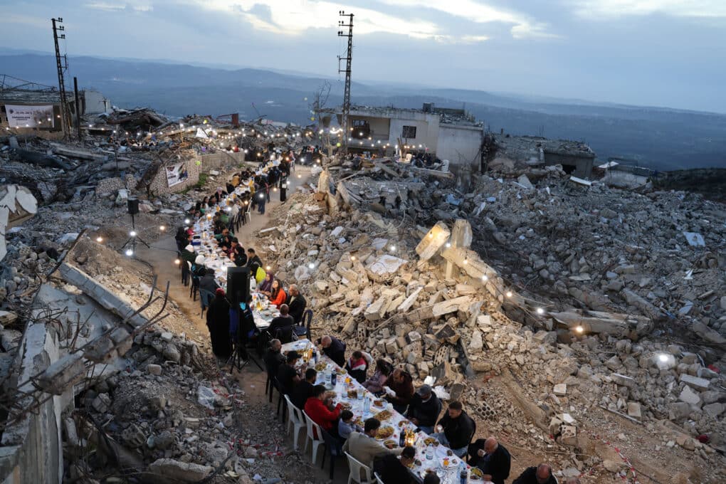 Color photograph of people seated at very long table with white cloth, set up on a winding road amidst fields of concrete rubble.