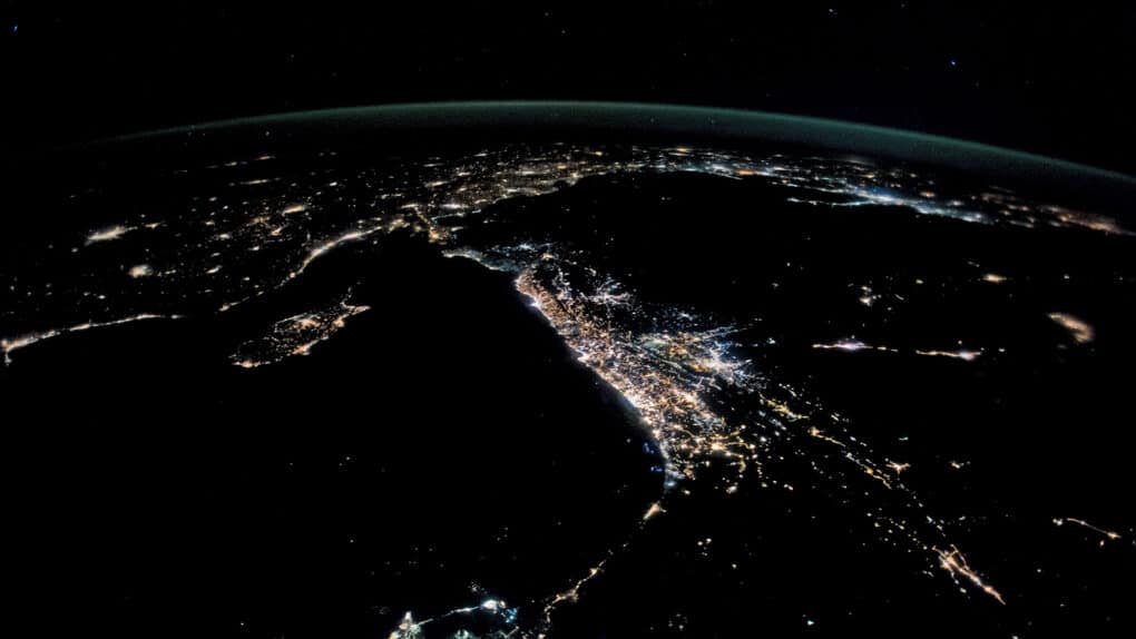 Aerial night photograph of narrow strip of land densely settled and brightly lit.