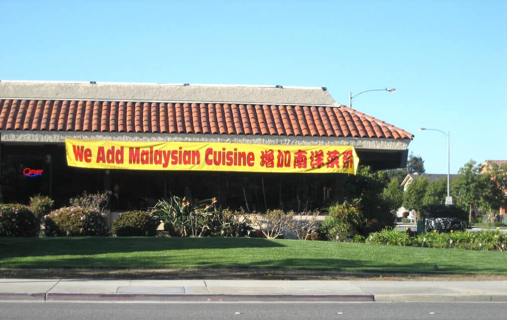 Color photo of restaurant with huge vinyl banner, bright yellow with red block letters, that says "We Add Malaysian Cuisine."