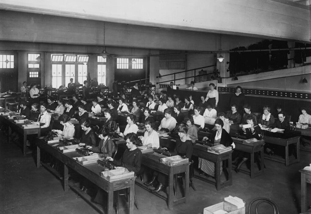 A black and white photo of about fifty women seated at desks in a large room, making calculations.