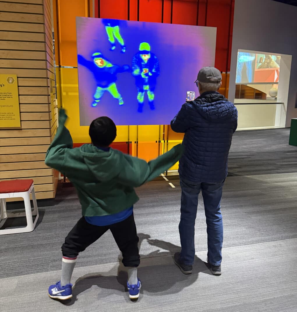 An older man and a young boy are watching a digital projection of themselves in infra-red video; the boy is dancing and the older man is taking a photo.