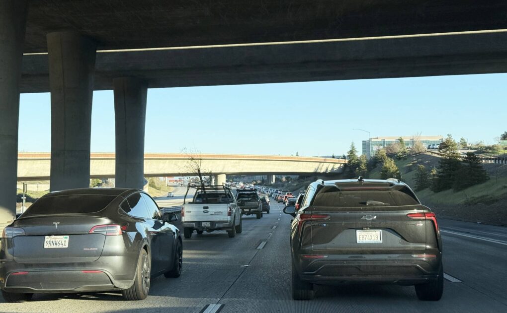 Mundane photo of highway filled with cars, apparently taken from a car, with a black Tesla in foreground.